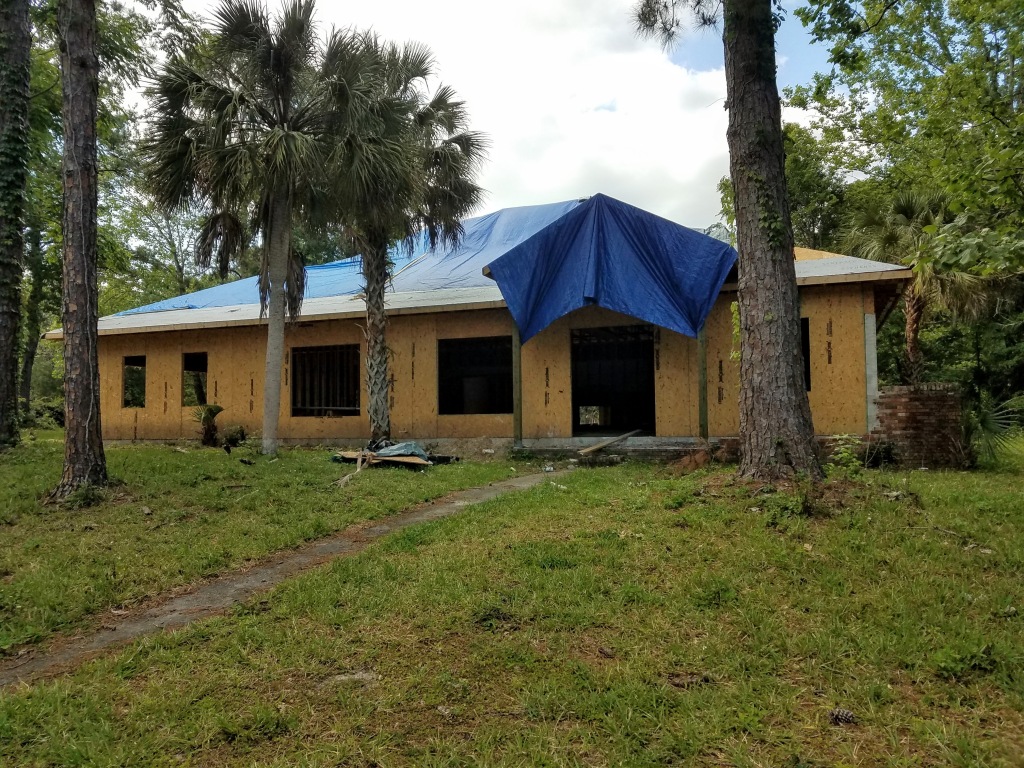 a seemingly abandoned house, partially constructed. The new wooden roof is covered with tarps, not shingles. The newly built walls leave gaping holes for windows and doors. The yard is vacant of any evidence that work on the house will continue.